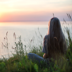 A woman is sat in the grass with her back to the viewer. She has long hair and is looking out to sea.